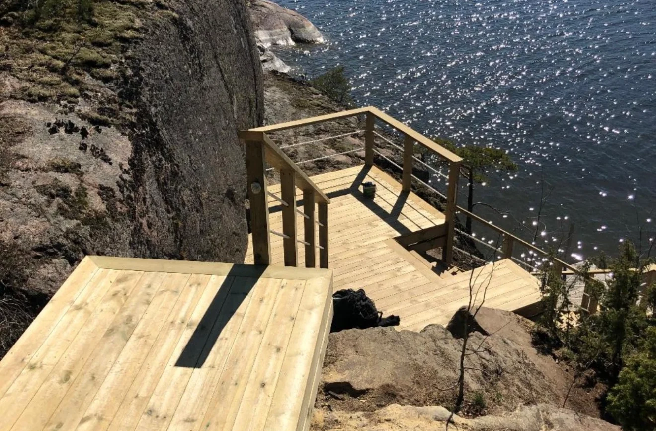 Nybyggt trädäck och trappa i ljust virke, stabilt förankrat på en brant berghäll i Stockholms skärgård.